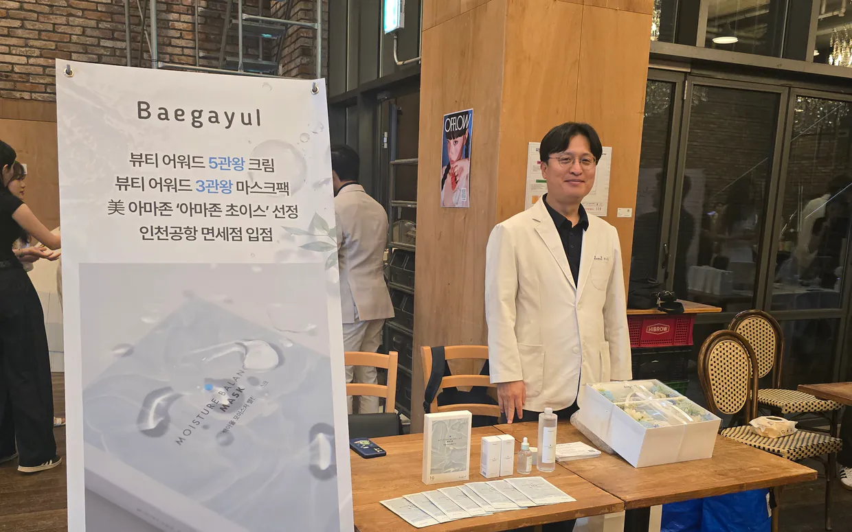 Dr. Jo Seok-yong standing beside a Baegayul display with the serum and sheet mask at a Korean beauty event
